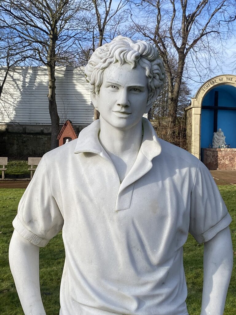 Statue of Saint Carlo Acutis, a young man in a polo shirt, at Carfin Grotto in Scotland.